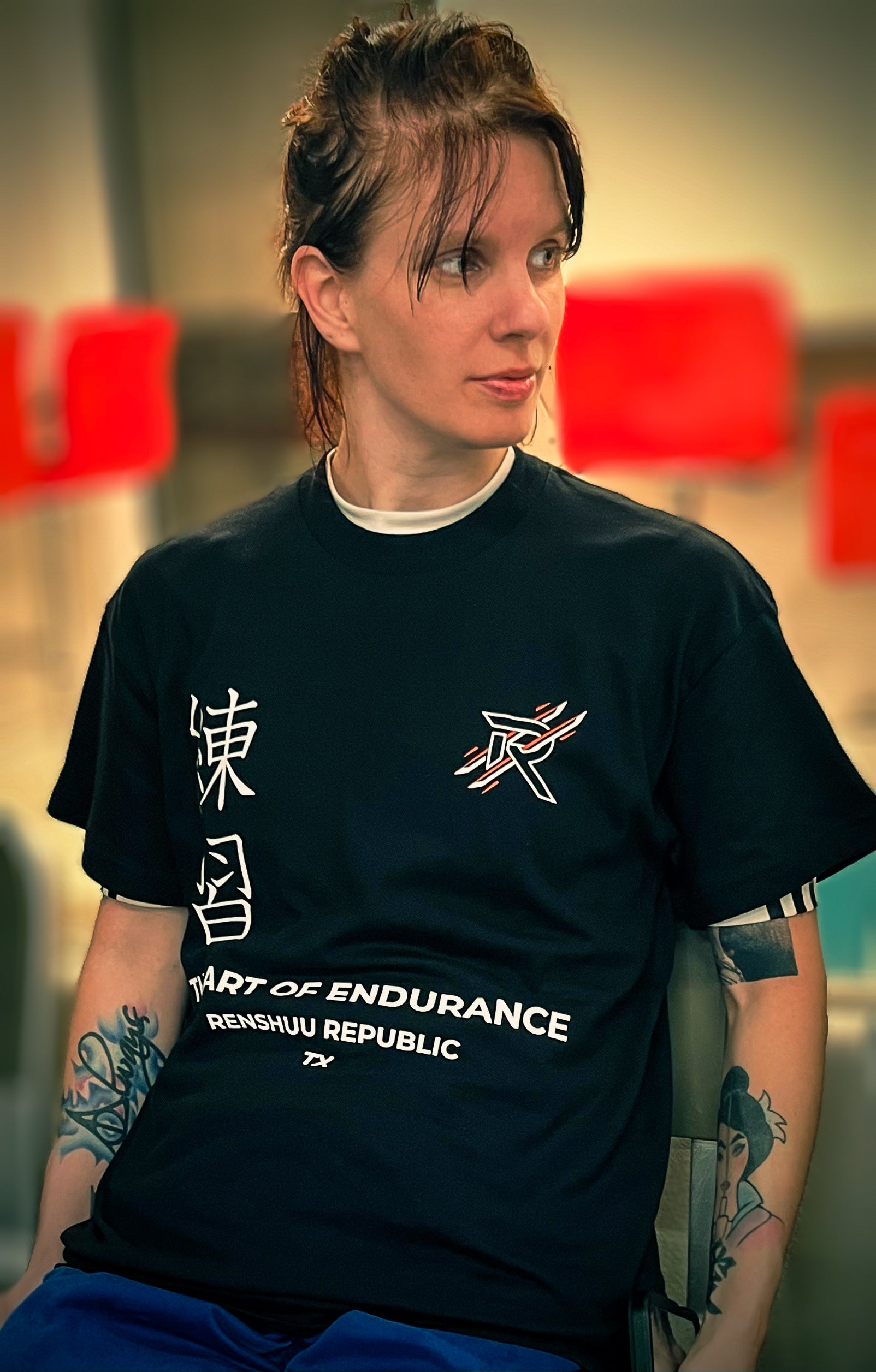 Person wearing a black t-shirt with text and graphics, sitting in a blurred indoor setting.