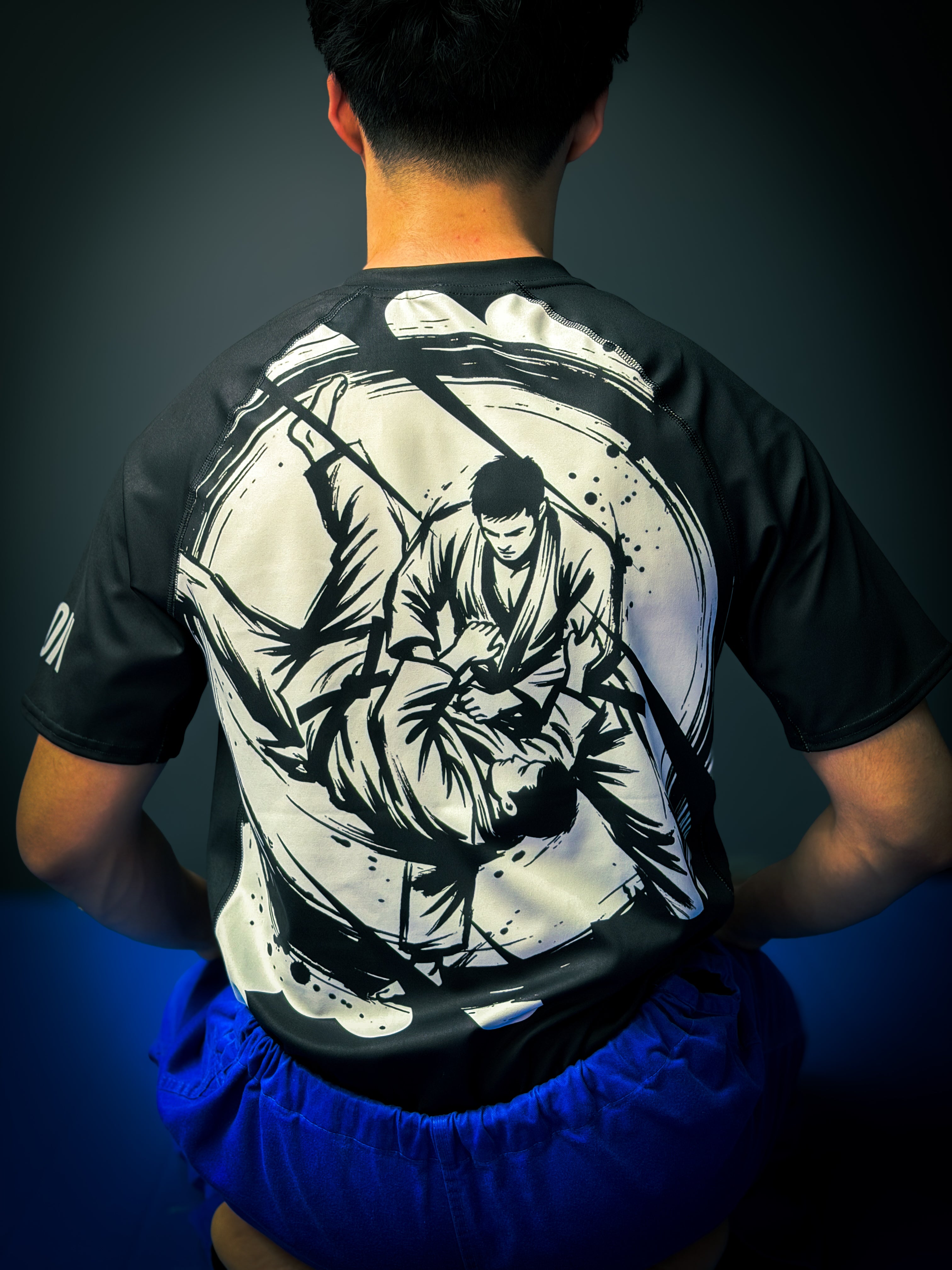 Person wearing a black t-shirt with a white graphic design on the back, standing against a dark background.