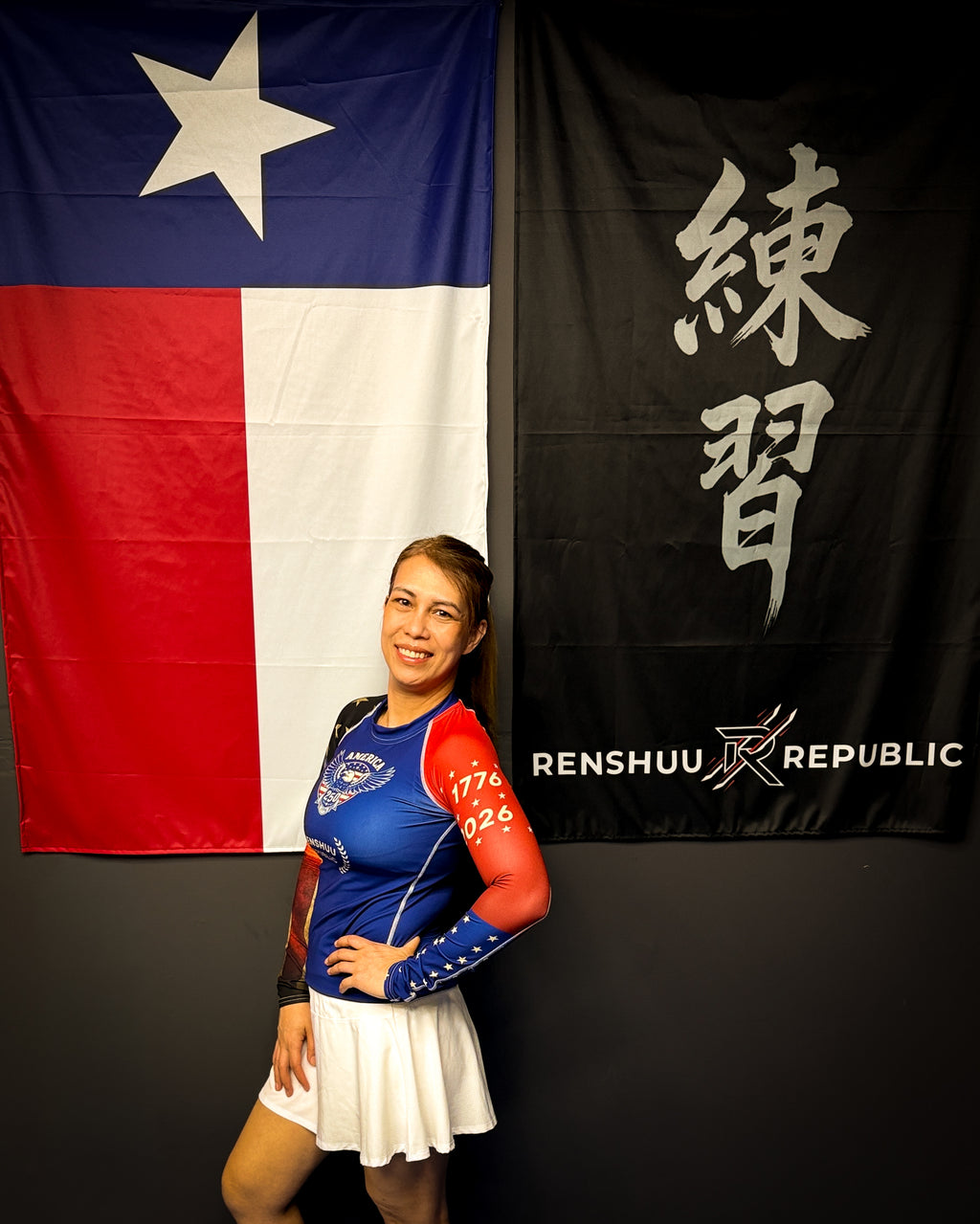 Person wearing a blue and red outfit with a white skirt, standing in front of a flag and a banner with text.