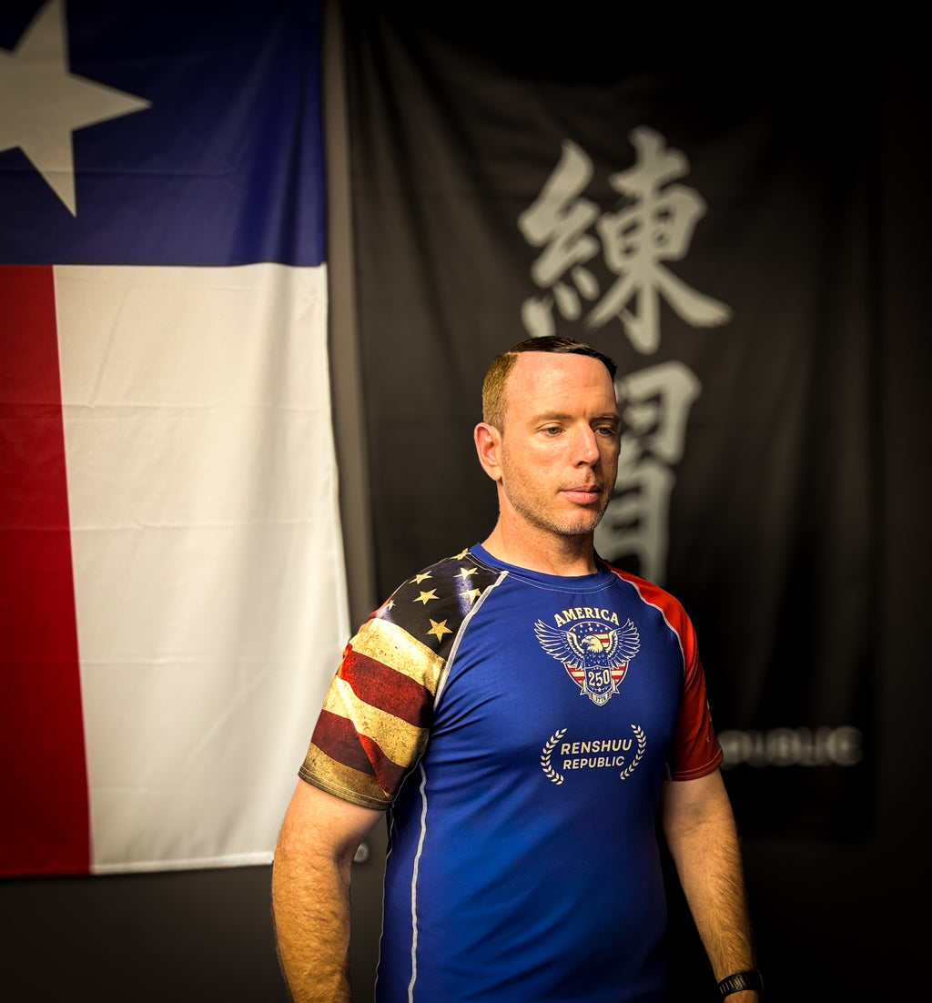 Man wearing a blue t-shirt with American flag design in front of a flag and black banner with white text.