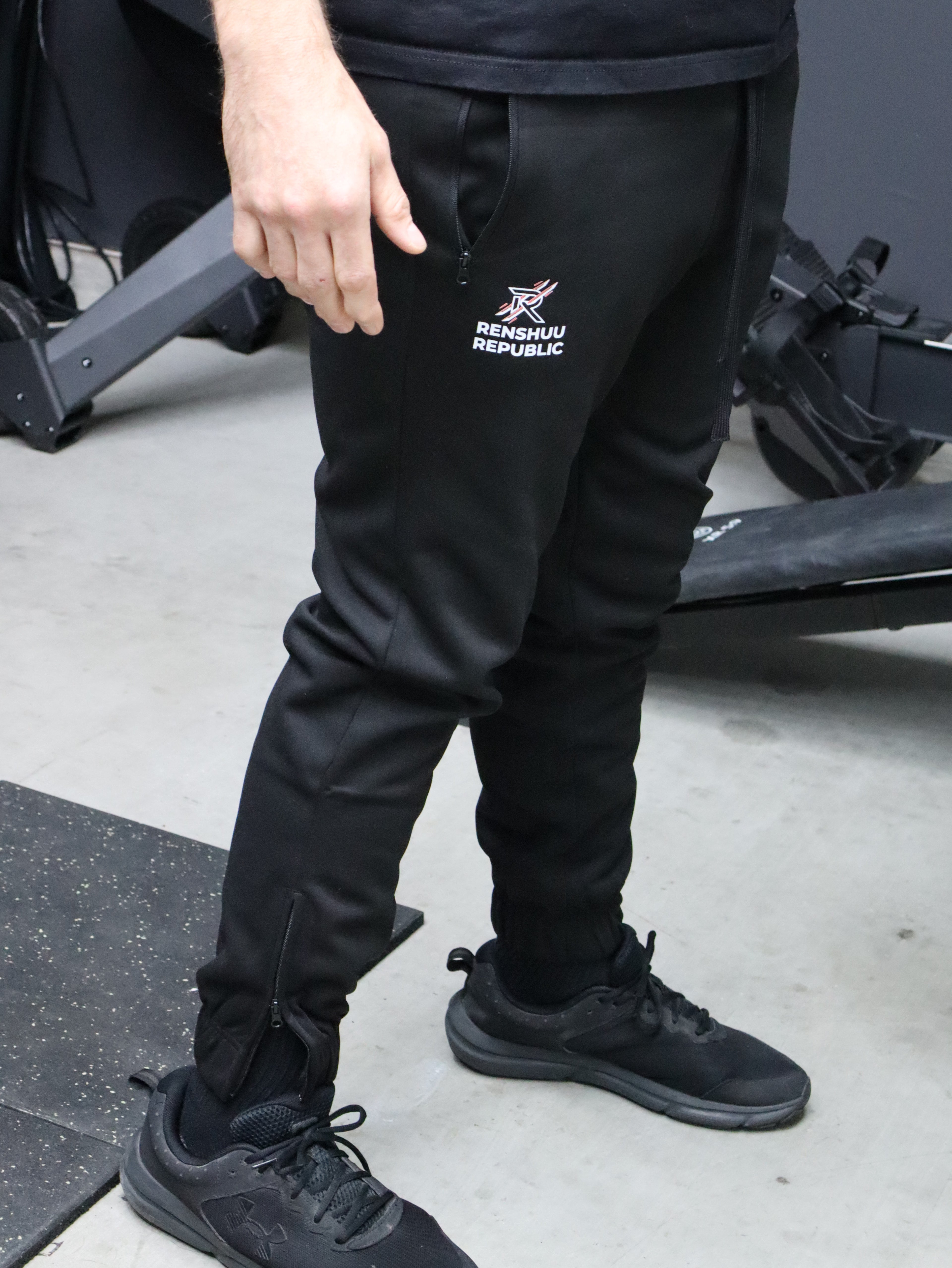 Person wearing black pants with a logo and black shoes on a gray floor.