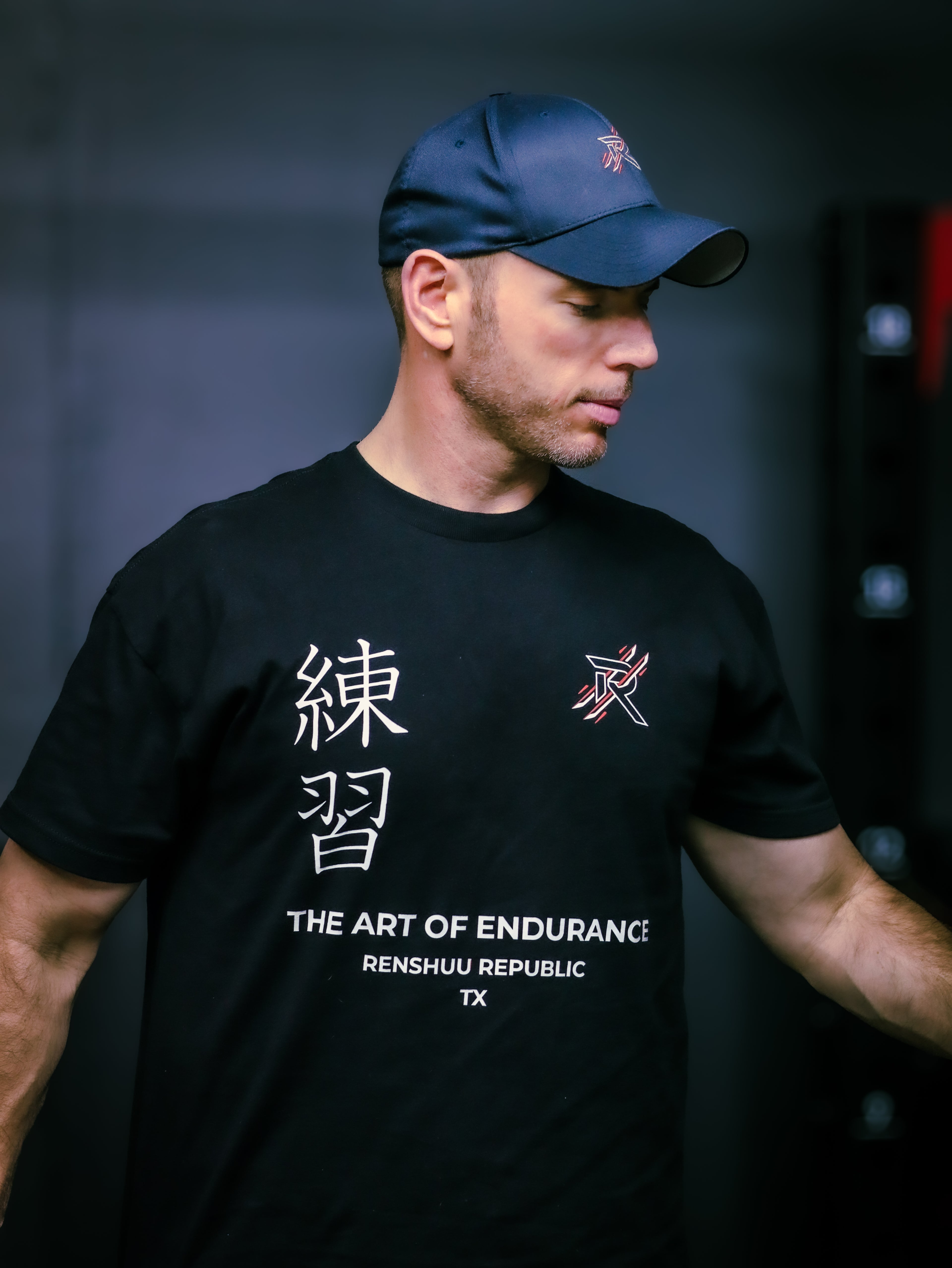 Man wearing a black t-shirt with text and logos, standing against a dark background