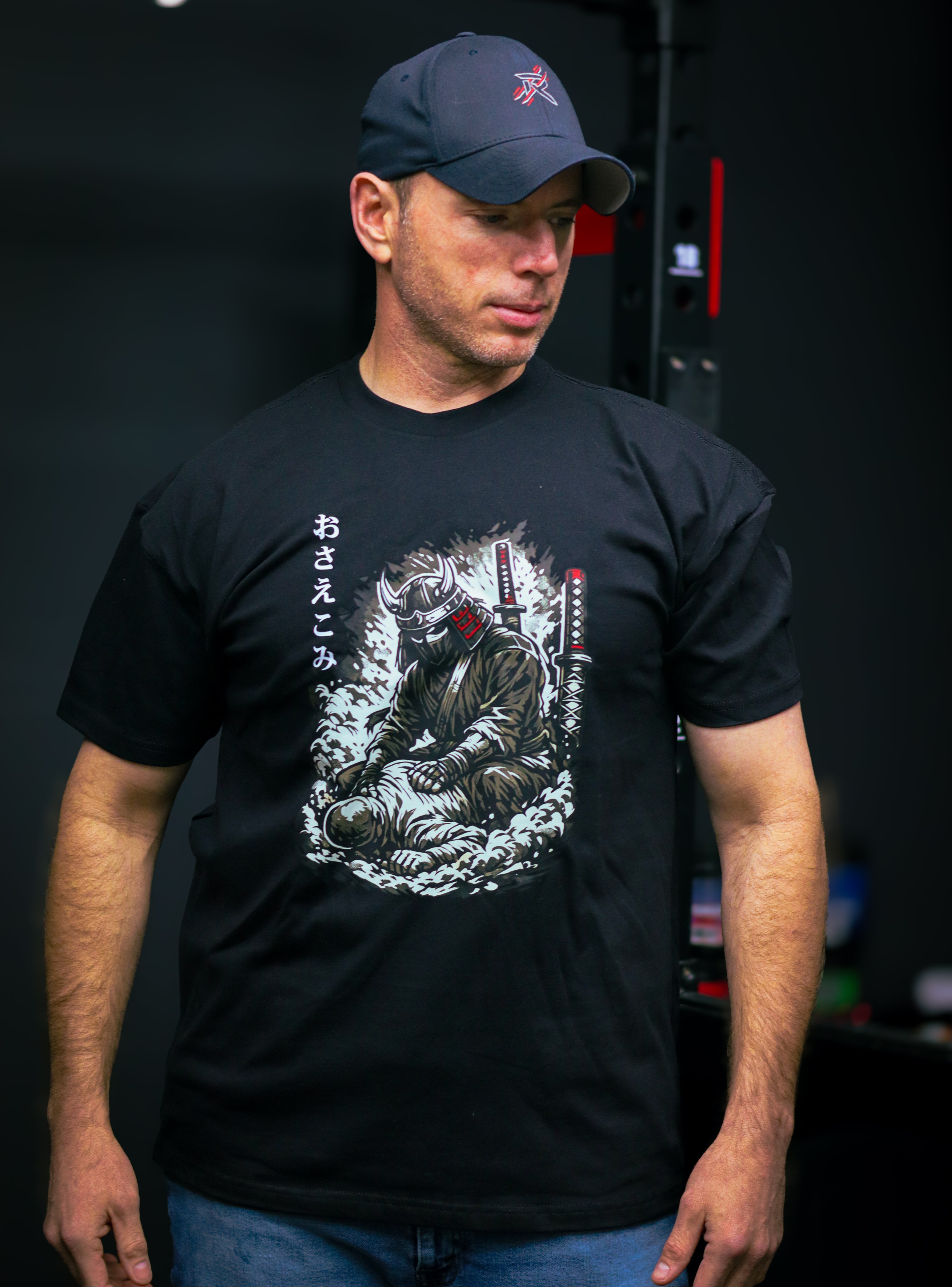Man wearing a black t-shirt with a graphic design and a cap, standing against a dark background.
